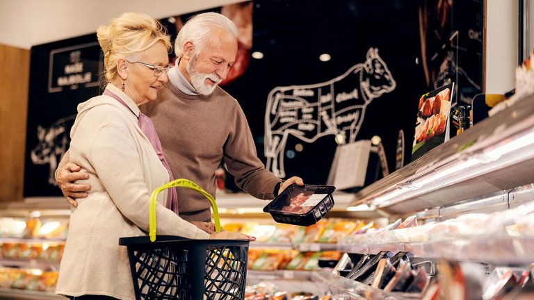 Ein älteres Paar steht im Supermarkt an der Fleisch-Selbstbedienungstheke. Der Mann hat seinen Arm um die Frau gelegt, in der anderen Hand hält er eine Packung Fleisch. Die Frau hat ihre Arme auf einem Einkaufskorb abgelegt. Sie lächeln und haben gute Laune.