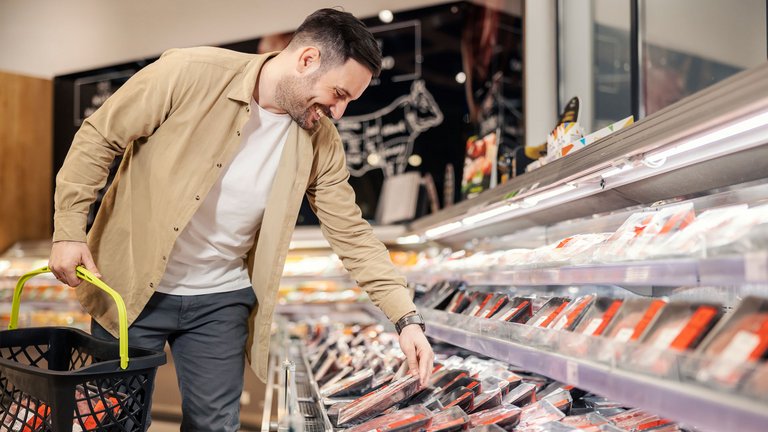 Ein junger Mann steht im Supermarkt an der Fleisch-Selbstbedienungstheke. Der Mann hat einen Einkaufskorb, in der anderen Hand hält er eine Packung Fleisch. Er lächelt und hat gute Laune.