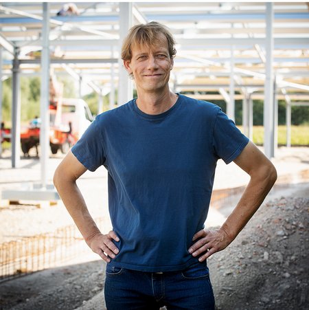 Ein Mann mit blonden, mittellangen Haaren und in einem blauen T-Shirt steht auf einer Baustelle. Er stemmt die Hände in die Hüften. Hinter ihm sieht man einen Haufen mit Dreck und Schutt, das Gerüst für ein großes Gebäude, ein Auto mit Hebebühne und Metallgitter für den Fußboden des Gebäudes.