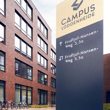 Im linken Teil des Bildes ist ein rotes Backsteingebäude. Es ist ein Bürogebäude mit vielen Fenstern. Im rechten Teil des Bildes steht ein Wegweiserschild. Der obere Teil des Schildes hat eine gelbliche Farbe, darauf steht "Campus Loddenheide." Der untere Teil des Schildes ist schwarz, darauf sieht man, in welche Richtung man gehen soll zu Fridthjof-Nansen Weg 3 / 3a oder zu Fridthjof-Nansen Weg 5 / 5a  